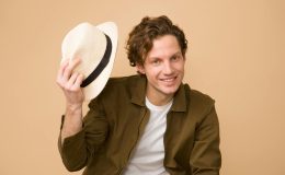 A cheerful man with curly hair tips his hat in a bright studio portrait.