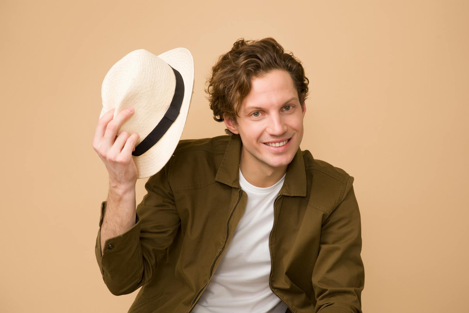 A cheerful man with curly hair tips his hat in a bright studio portrait.