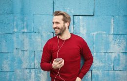 A joyful man in a red sweater smiles while listening to music on his smartphone against a blue brick wall.