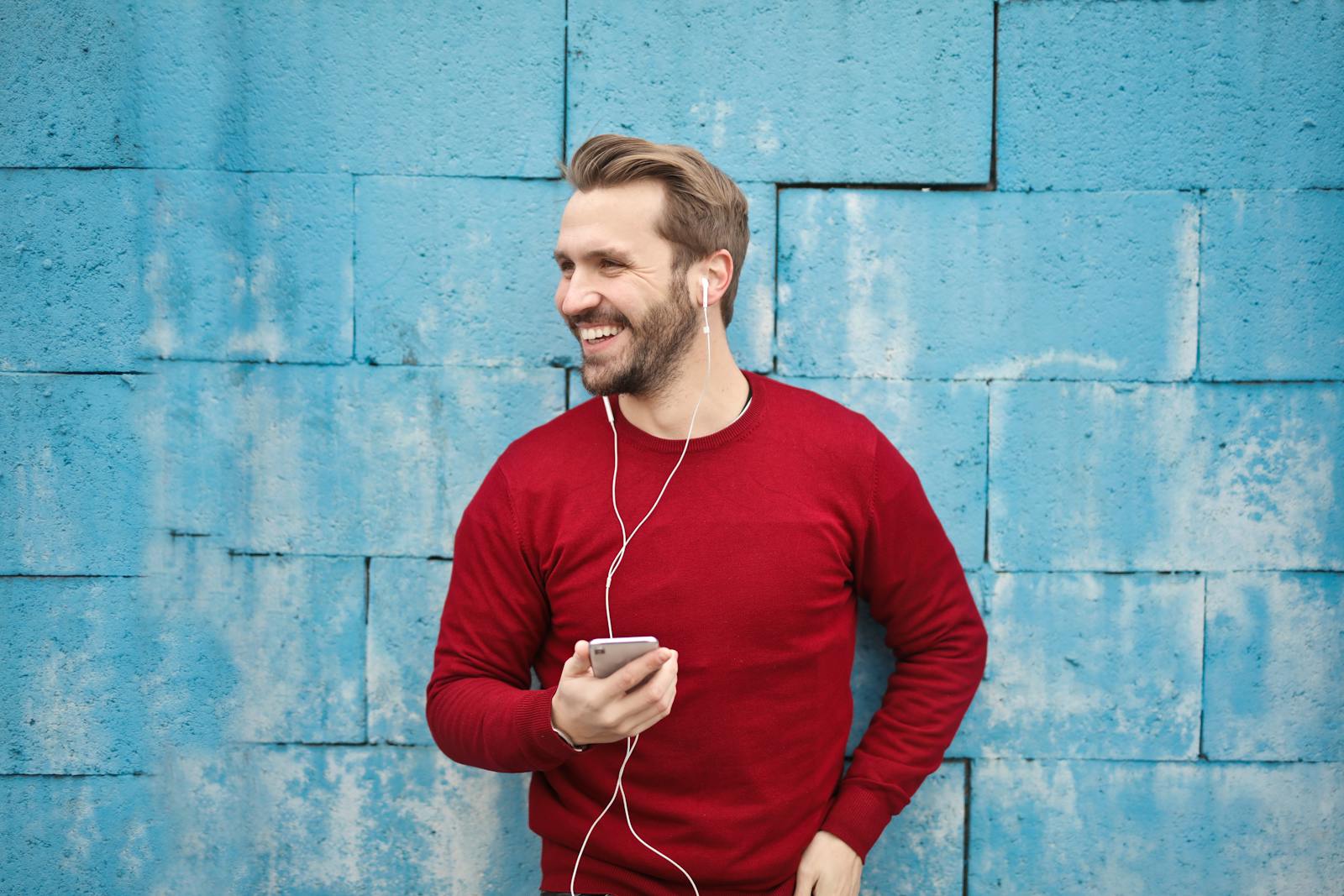 A joyful man in a red sweater smiles while listening to music on his smartphone against a blue brick wall.